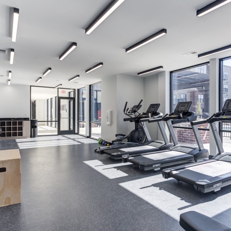 Modern fitness center with treadmills, large windows, dark rubber flooring, and LED lighting at Station 250 apartment amenities Mundelein IL